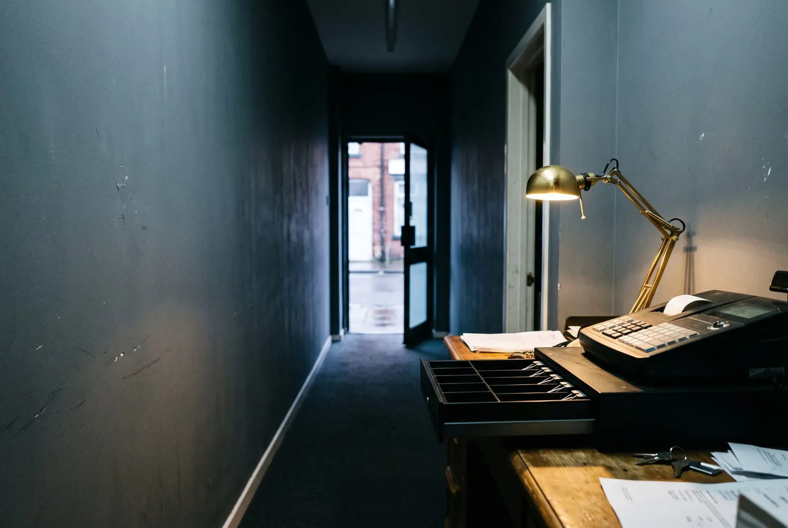 An empty shop back office at night with the street door open at the end of the corridor and the till drawer standing open, illustrating that AI agents can now pay merchants who have built nothing for them.