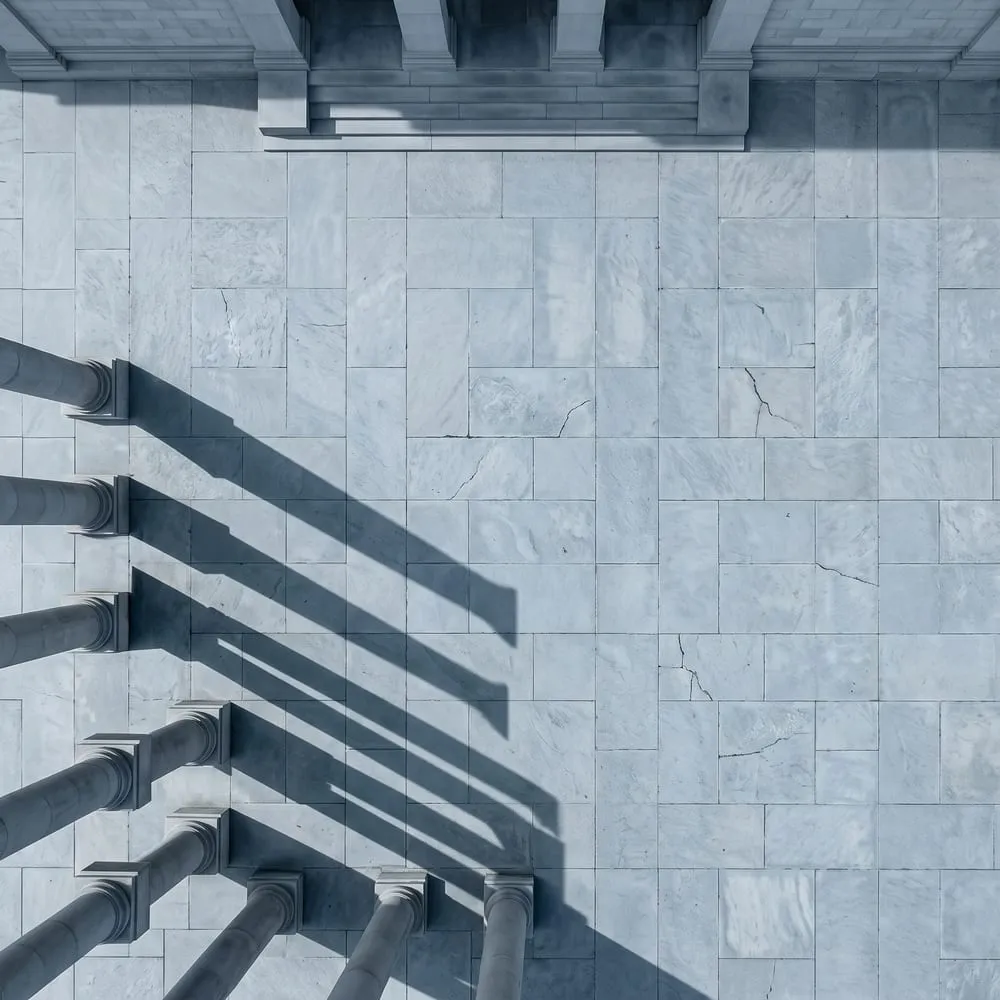 An empty courthouse forecourt from directly above at dawn with long column shadows, illustrating the unresolved legal position between Amazon and Meta over shopping agents.